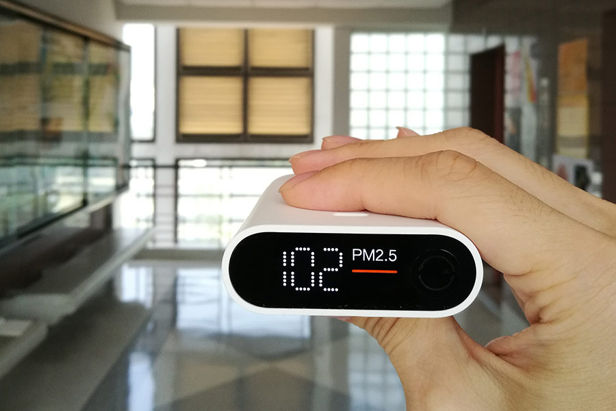 Hand holding a PM2.5 air quality monitor displaying "102"